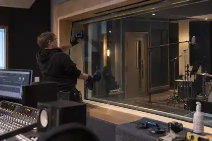 Recording studio interior from control desk: technician glazing a large angled acoustic observation screen within deep timber jambs, dark acoustic panels and microphone booth beyond.