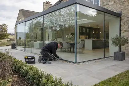 Architectural exterior hero: finished contemporary glass extension on a stone British house, full-height structural glazing with slim supports, a technician checking the junction at ground level, wide three-quarter view.