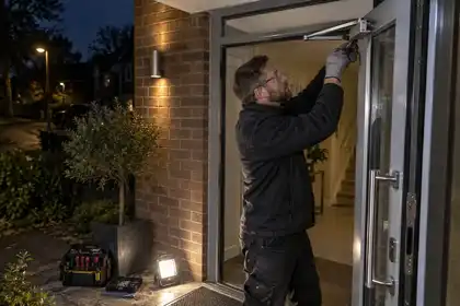 After-dark residential porch: professional repairing a damaged aluminium door closer bracket while the glazed entrance door is supported open, battery lamp on floor, tools neatly arranged, no forced-entry activity.